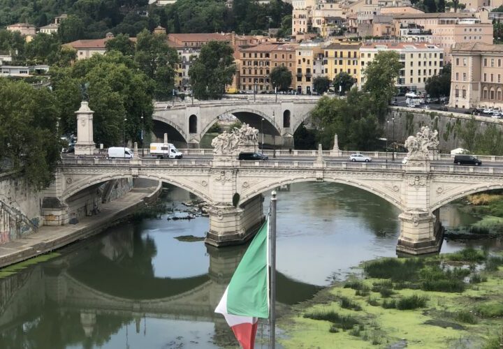 Eine befahrene Brücke führt über einen großen Fluß. Im Hintergrund sind viele Gebäude, vorne ist die italienische Flagge zu sehen.