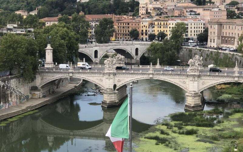 Eine befahrene Brücke führt über einen großen Fluß. Im Hintergrund sind viele Gebäude, vorne ist die italienische Flagge zu sehen.