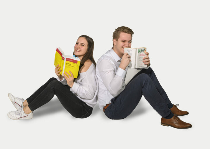 Eine junge Frau und ein junger Mann sitzen Rücken an Rücken. Sie lachen und halten jeder ein aufgeklapptes Buch in der Hand. Sie tragen schwarze Hose und weißes Hemd.