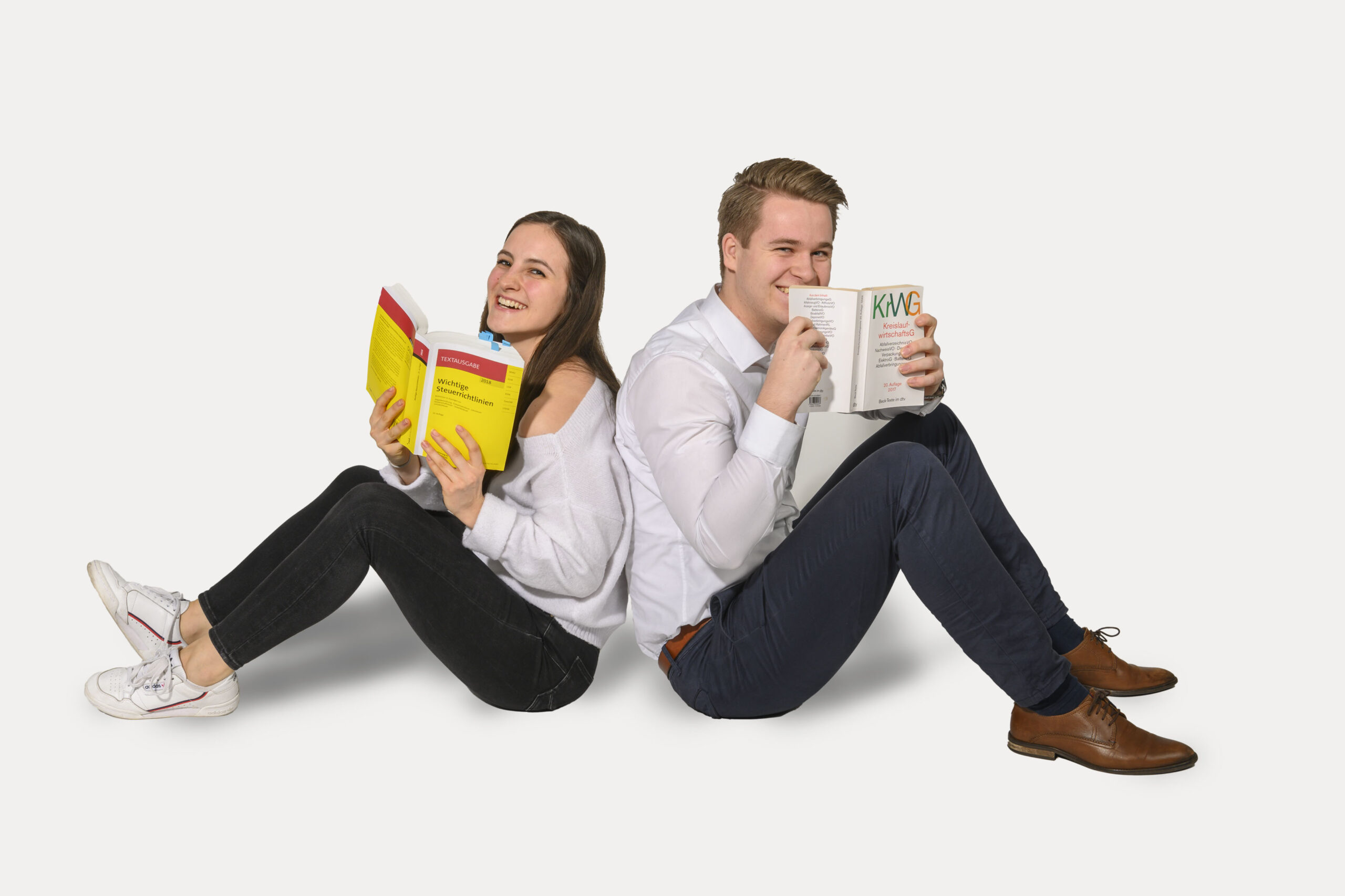 Eine junge Frau und ein junger Mann sitzen Rücken an Rücken. Sie lachen und halten jeder ein aufgeklapptes Buch in der Hand. Sie tragen schwarze Hose und weißes Hemd.