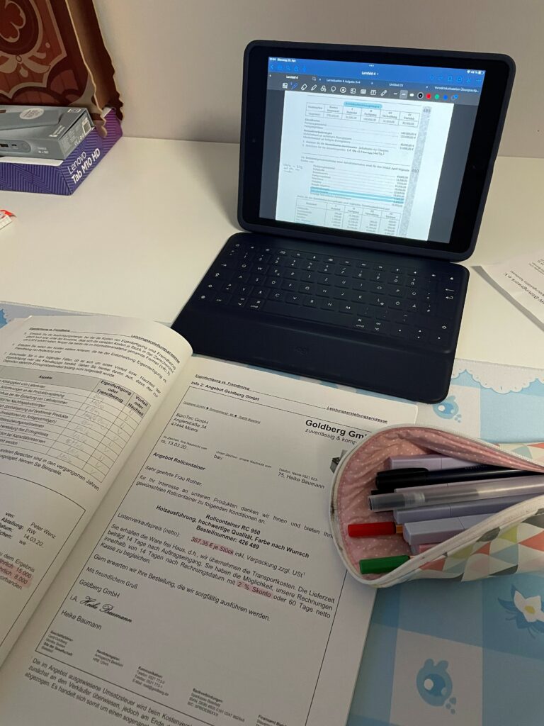 Ein Laptop auf einem Tisch. Darunter liegt ein geöffnetes Heft und ein Mäppchen mit Stiften.