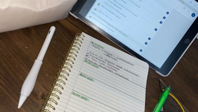 Ein Tablet liegt auf einem Tisch, darunter ein Notizblock. Daneben befinden sich ein weißes Mäppchen, ein Digitalstift, mehrere Stifte und ein Geodreieck.