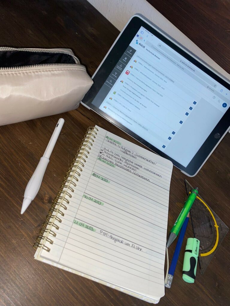 Ein Tablet liegt auf einem Tisch, darunter ein Notizblock. Daneben befinden sich ein weißes Mäppchen, ein Digitalstift, mehrere Stifte und ein Geodreieck.