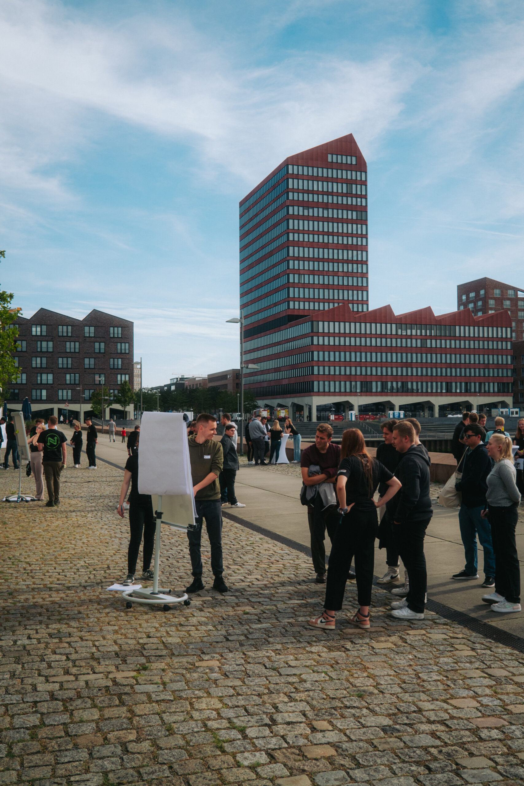 Mehrere junge Erwachsene stehen in kleinen Gruppen auf einer gepflasterten Straße und arbeiten an einem großen Klemmbrett auf einem rollbaren Ständer. Im Hintergrund stehen weitere Personen an einem Stehtisch sowie moderne Gebäude.